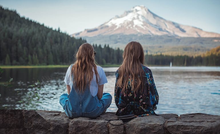 two women sitting on rock facing on body of water and mountain